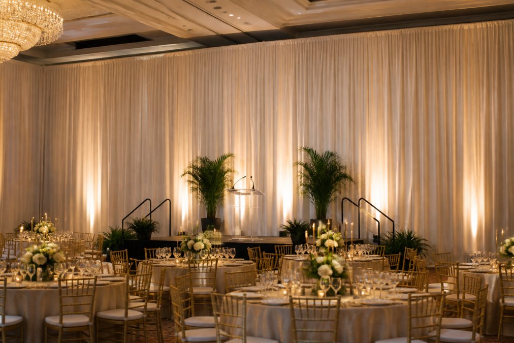 Elegant corporate gala ballroom with floor-to-ceiling ivory pipe and drape fabric creating a dramatic backdrop, warm uplighting washing the drapes, and a branded stage in the foreground