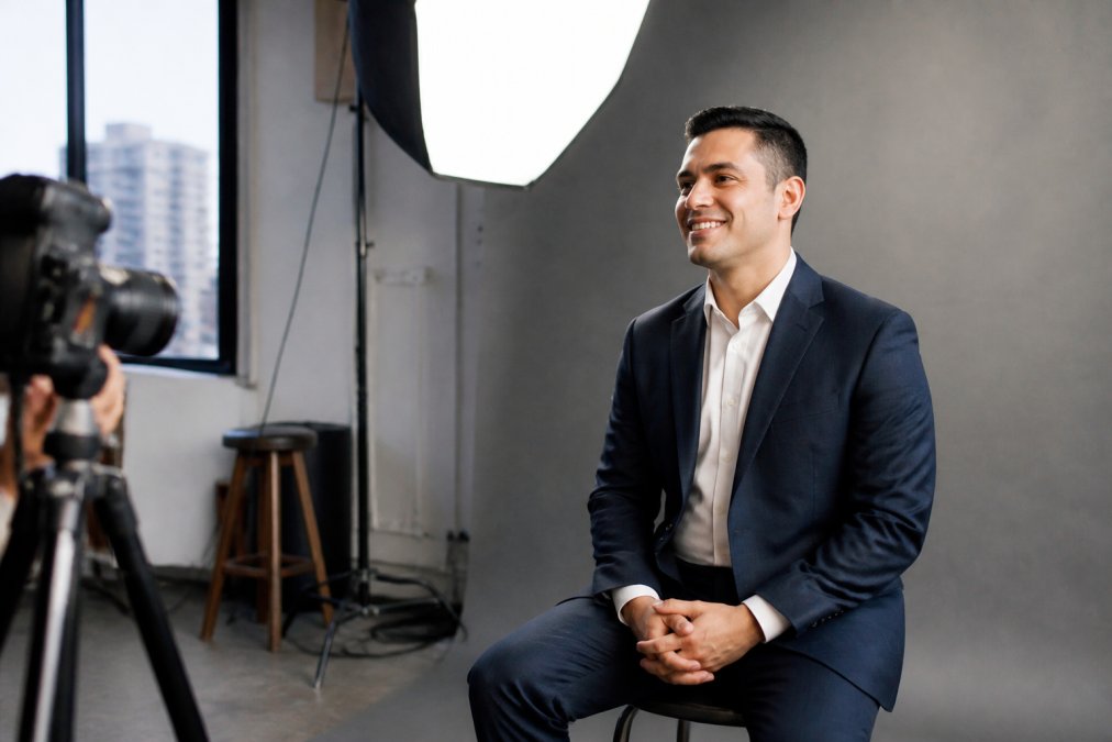 Professional in business attire sitting in a modern studio with soft directional lighting, displaying confident posture and natural expression during a corporate headshot session