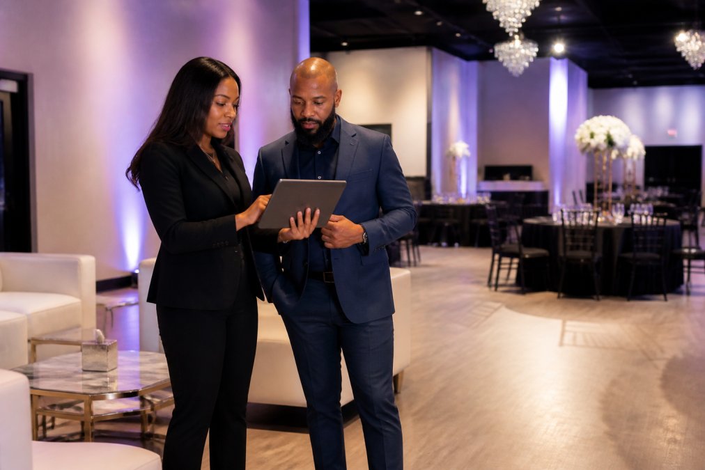 A corporate event planner and team leader touring an elegant modern event venue with lounge furniture and soft up-lighting, discussing layout options on a tablet