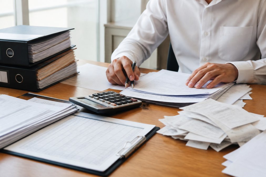 Professional accountant organizing corporate tax documents, financial statements, and receipts on a wooden desk with calculator and filing folders in natural office lighting