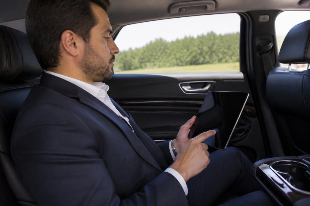 Business professional in car reviewing documents on tablet with privacy screen protector clearly visible, showing secure work environment during transit, natural light from window