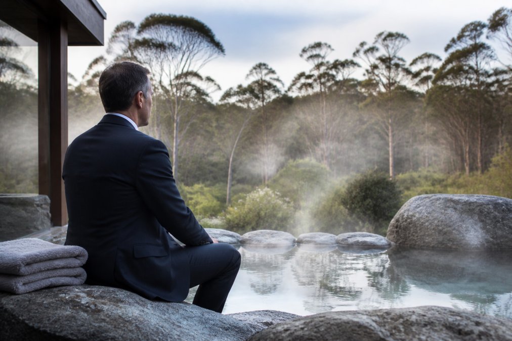 Senior executive in professional attire sitting peacefully by a natural thermal pool surrounded by native forest and soft morning mist, with serene water reflections and natural stone features