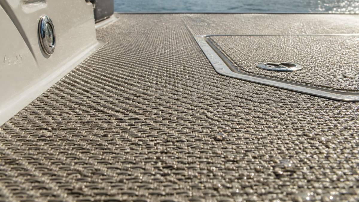 Close-up of woven vinyl marine flooring installed on a boat deck in bright sunlight, showing textured surface with water droplets beading off, demonstrating water resistance and durability