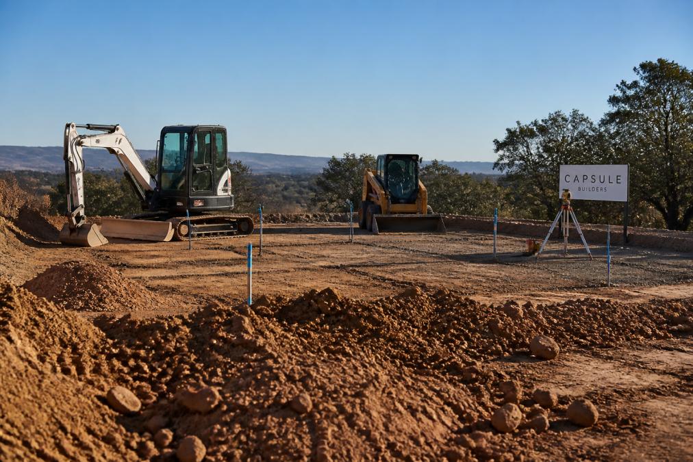 Construction site with excavation equipment and machinery preparing land, showing cleared earth, surveying markers, and foundation preparation on a regional property with natural landscape