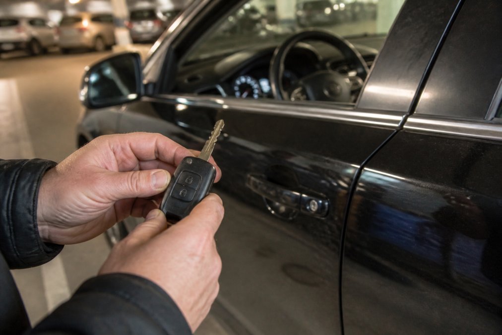 Close-up of a person's hands holding a black car transponder key fob next to a vehicle door handle in a parking garage, with the car's ignition visible in the soft background lighting