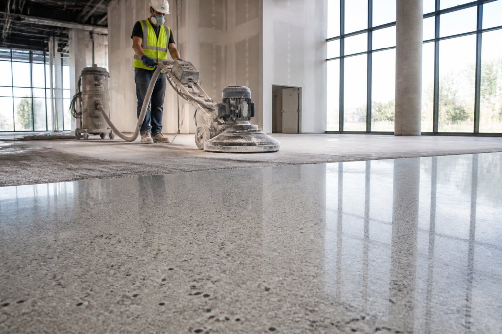 A flooring contractor in safety gear operating a heavy-duty terrazzo grinding machine on a large commercial job site, with a polished floor section visible in the foreground