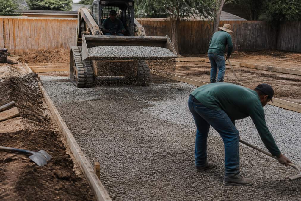 Hardscape crew performing excavation and sub-base preparation for patio installation, with heavy equipment visible and workers installing base aggregate layers, natural daylight on active construction site