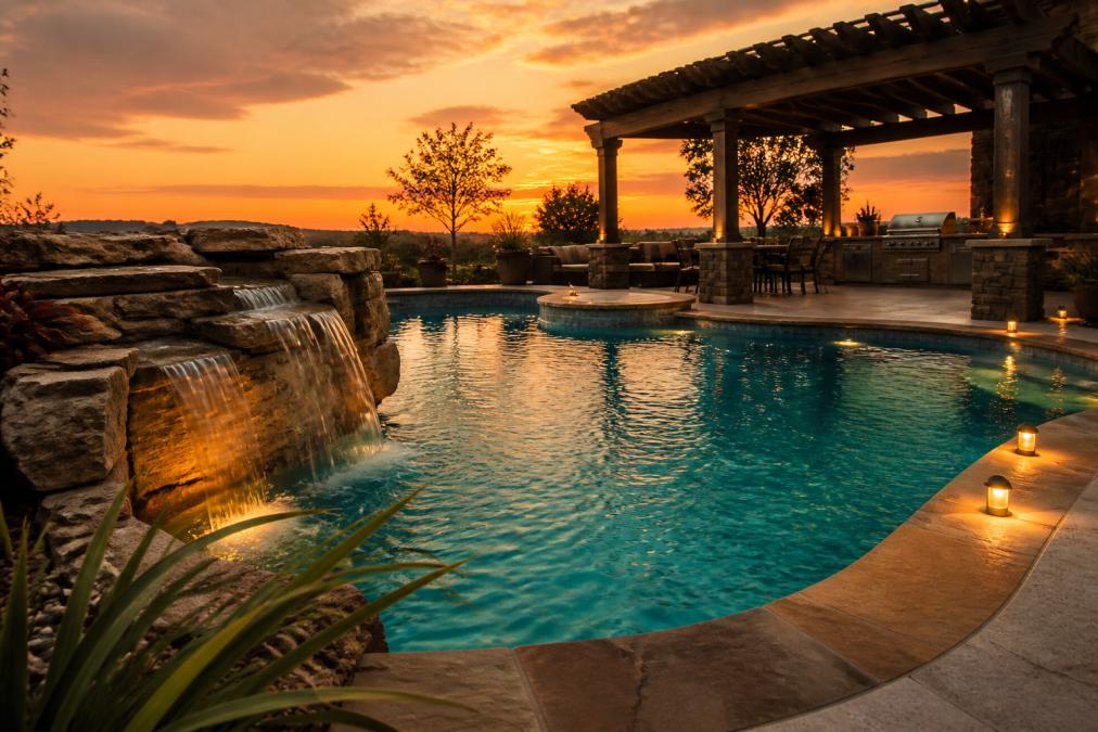 A luxury custom pool with a natural stone waterfall feature cascading into turquoise water, LED lighting glowing along the pool edges at golden hour, with a modern outdoor kitchen and pergola structure visible in the background against a warm sunset sky