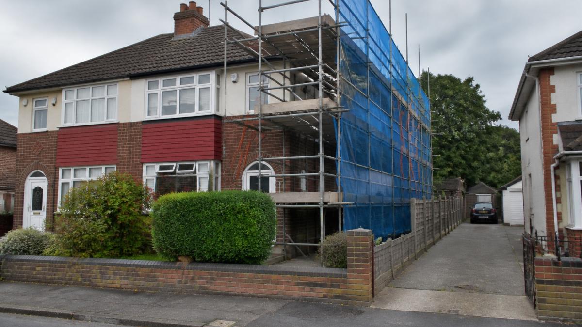 A fixed scaffold erected along the side of a typical semi-detached house, showing the full elevation with protective netting and a ladder access point, residential street setting