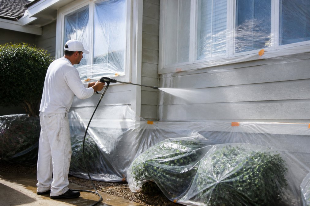 Professional painter using pressure washing equipment against house siding with water spray, protective coverings on landscaping and windows, bright daylight conditions