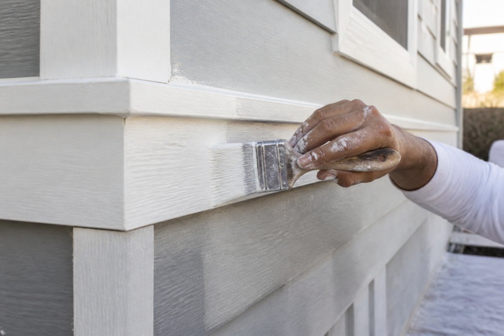 Painter's hands applying primer to wood trim and siding with brush, showing quality paint application on different surface types, natural outdoor lighting