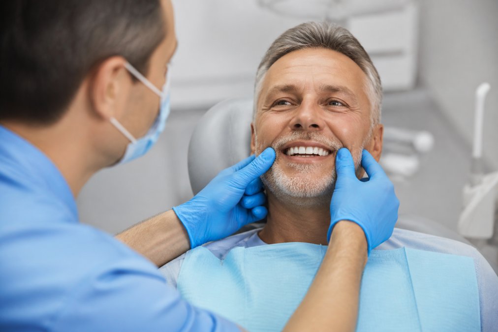 Close-up of a patient smiling confidently in a dental chair while a dentist in gloves gently examines their restored teeth, bright clinical setting with modern equipment visible