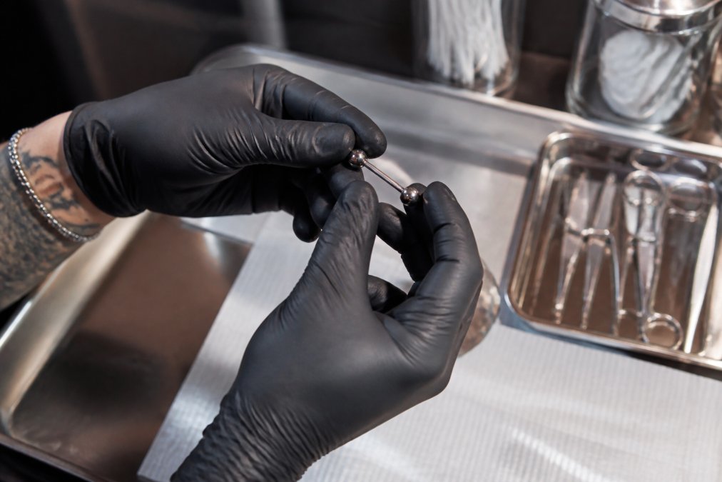 A close-up of a piercer's gloved hands holding a small, polished titanium barbell under bright studio lighting, with a sterile jewelry tray and tool kit visible in the background