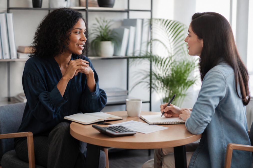 A coach and client sitting across from each other in a bright, modern office space, engaged in a one-on-one conversation with notebooks and coffee on the table