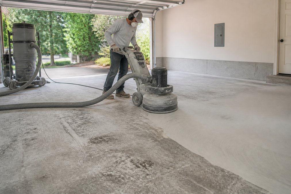A worker in safety gear using a concrete grinder on a residential garage floor, with dust extraction equipment visible and a half-prepped surface showing the contrast between rough and smooth concrete