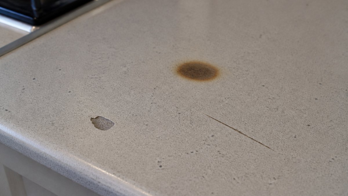 Close-up of a kitchen worktop showing various types of cosmetic damage including a burn mark, a chip, and a scratch, with natural kitchen lighting