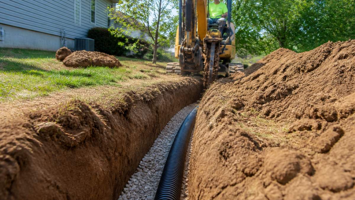 Professional excavation crew operating trenching equipment in residential yard with visible soil layers and drainage trench preparation
