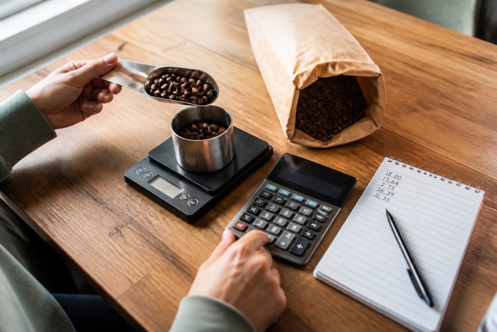 A person's hands using a digital kitchen scale to measure whole coffee beans next to a calculator, a bag of beans, and a notepad with numbers on a wooden kitchen counter