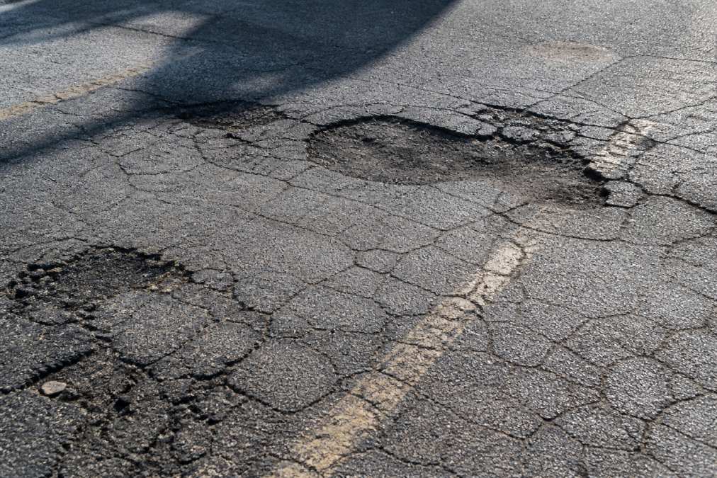 Close-up of asphalt surface showing extensive alligator cracking patterns, dark potholes, and faded striping with natural sunlight casting shadows across deteriorated pavement sections