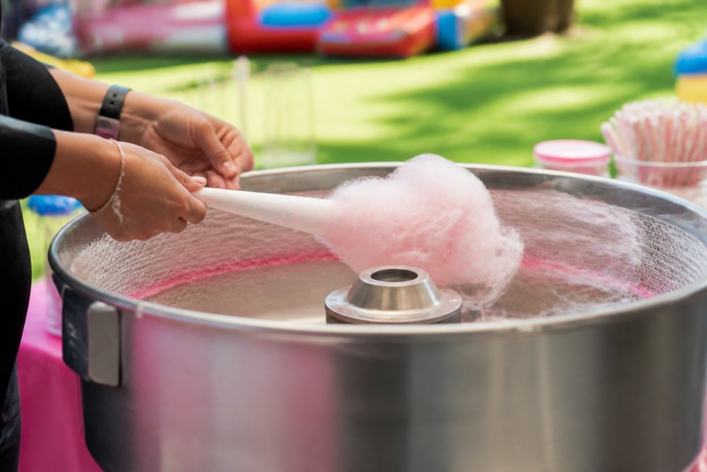 Close-up of a person's hands holding a paper cone under the spinning head of a cotton candy machine, collecting fluffy pink sugar floss at a bright outdoor party