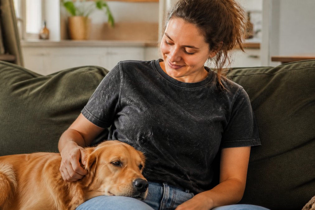 A woman in a casual graphic tee sitting on a couch with a golden retriever resting its head on her lap, visible pet hair on the fabric of the shirt, warm natural light, candid home setting