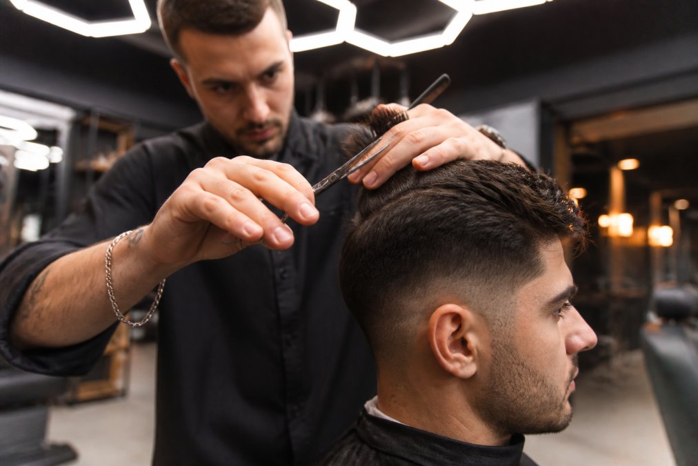 Close-up of a barber's hands using scissors to texture the top section of a man's thick dark hair, client seated in a leather chair, warm ambient light reflecting off hexagonal ceiling fixtures in an upscale barbershop