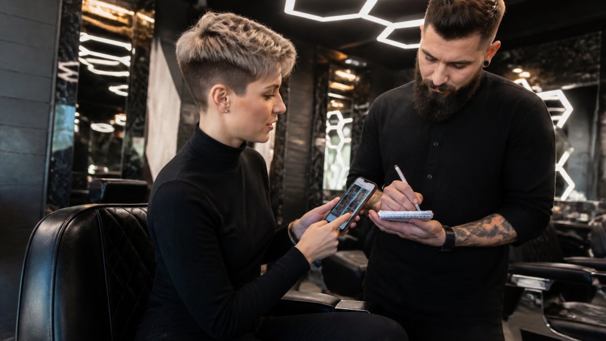 Une femme assise dans un fauteuil de barbier haut de gamme, montrant des photos de coupes courtes sur son téléphone à un barbier qui prend des notes, éclairage hexagonal en arrière-plan