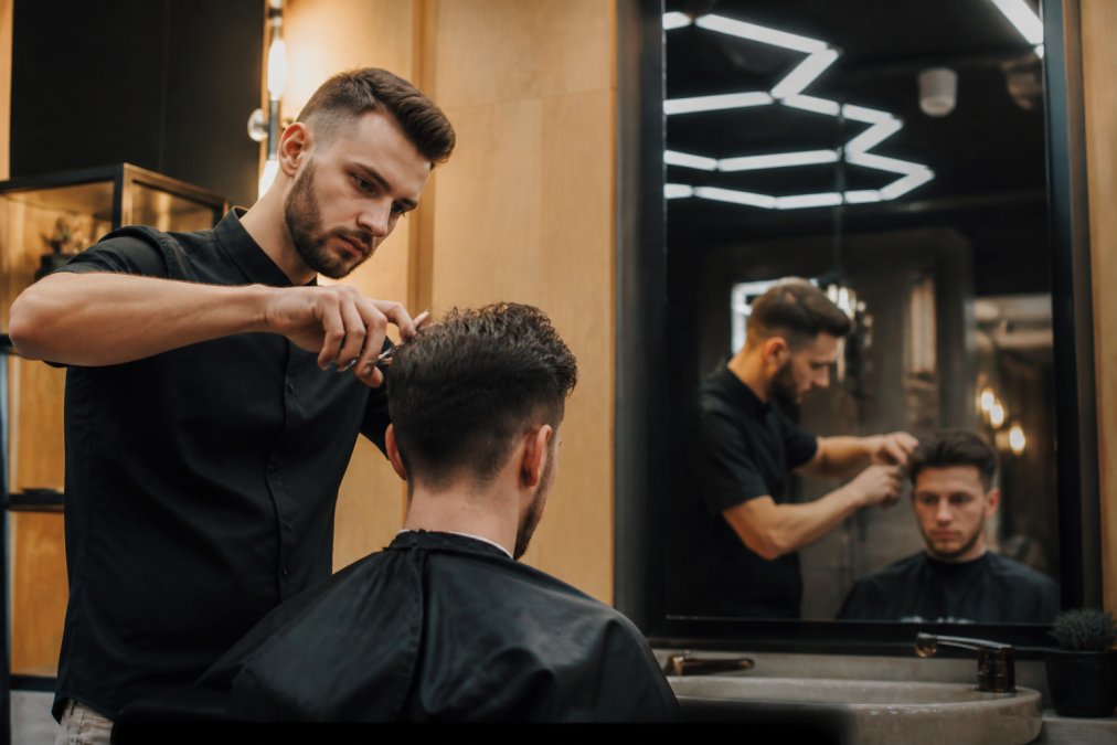 Un barbier concentré qui sectionne les cheveux d'un client aux ciseaux dans un salon moderne et éclairé, reflet dans un miroir, ambiance soignée et professionnelle
