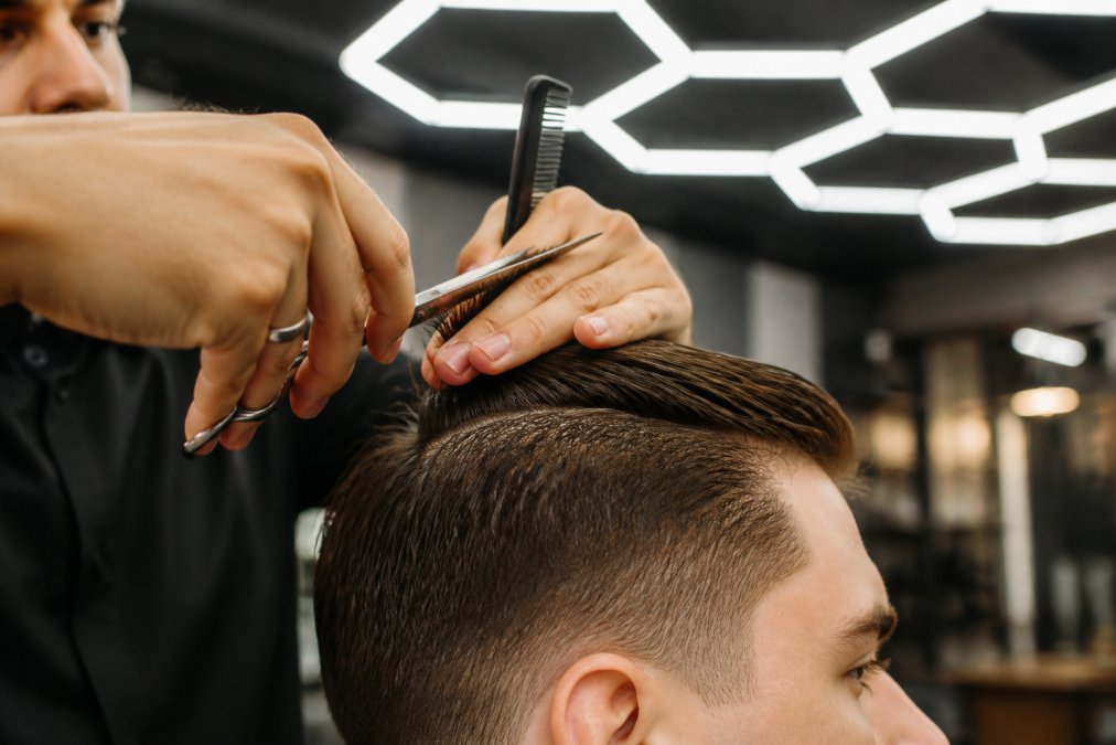 Gros plan sur les mains d'un barbier tenant des ciseaux de coiffure et un peigne professionnel, coupant une mèche horizontale sur le dessus du crâne d'un client, dans un salon éclairé par un éclairage hexagonal