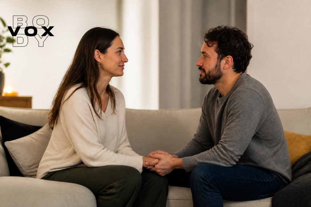 Two partners sitting face to face on a couch in a warm, softly lit room, holding hands and making gentle eye contact during what appears to be a guided conversation exercise