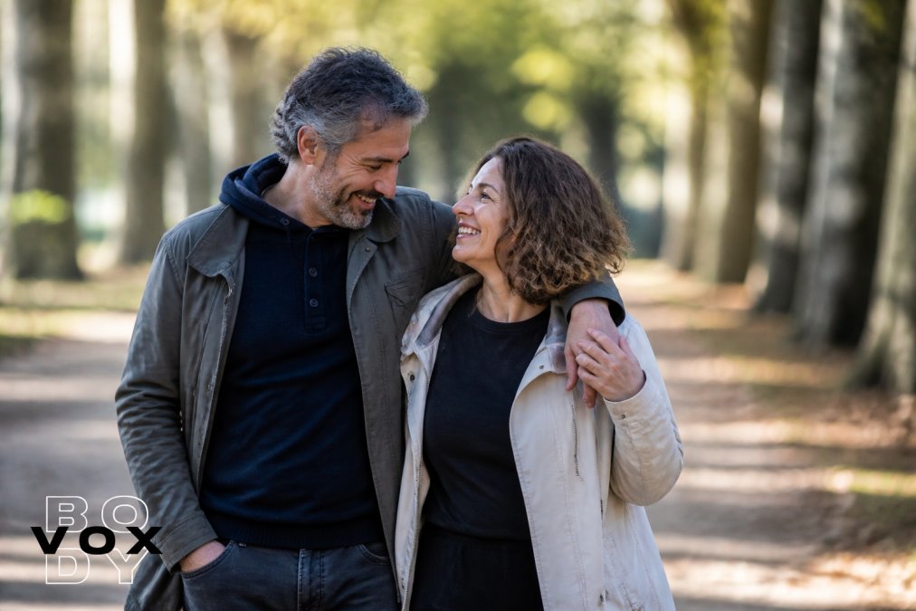 A couple in their mid-40s walking together outdoors on a tree-lined path in soft afternoon light, smiling and leaning into each other, conveying ease and renewed closeness