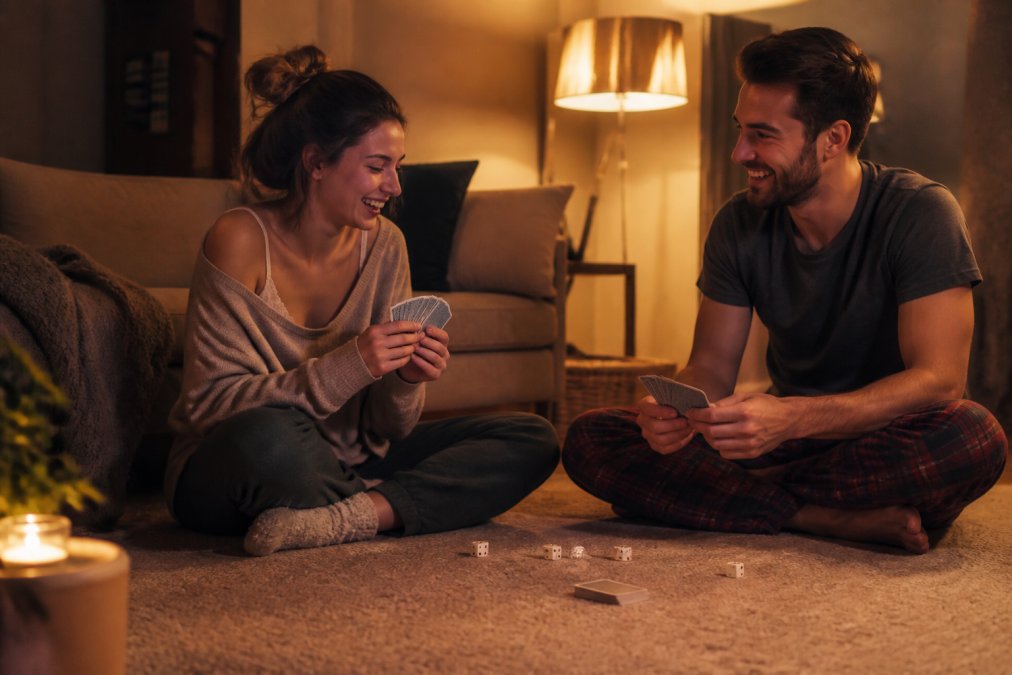 A couple sitting cross-legged on a living room floor at night, soft lamp light, playing a small card game with dice scattered nearby, both laughing comfortably in casual clothes