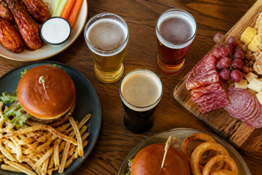 Overhead view of craft beer glasses filled with different styles, pale ale, amber, stout, arranged on a wooden table with plates of burgers, crispy fried chicken wings, and cheese and charcuterie boards nearby, natural afternoon lighting
