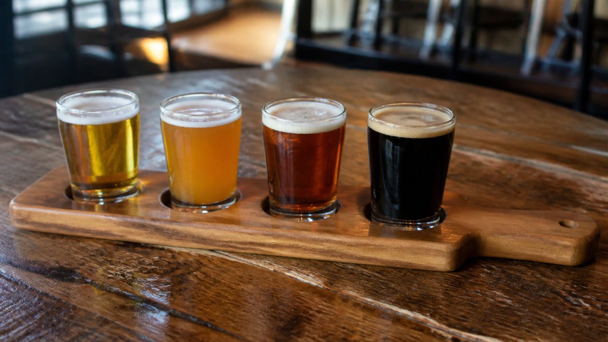 A wooden flight paddle holding four small beer glasses arranged from light golden lager to dark stout, set on a rustic taproom table with soft natural light