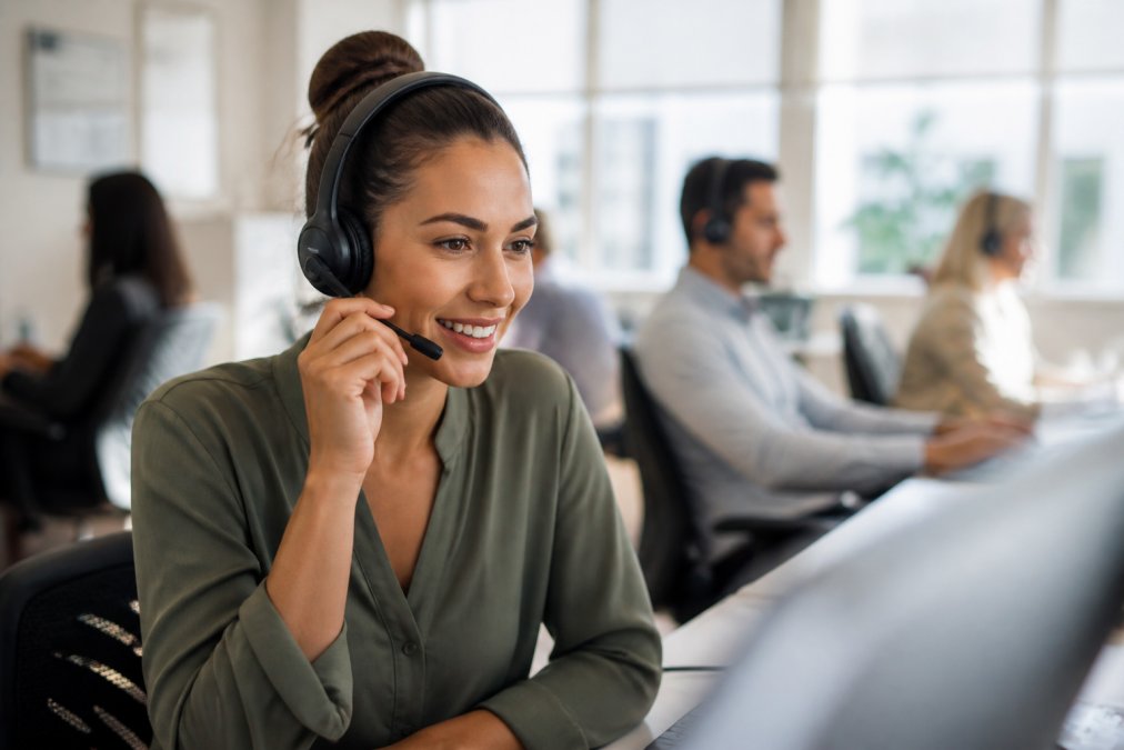 Customer service representative at a call centre desk wearing a headset, assisting a caller with a warm expression, modern office environment with natural light
