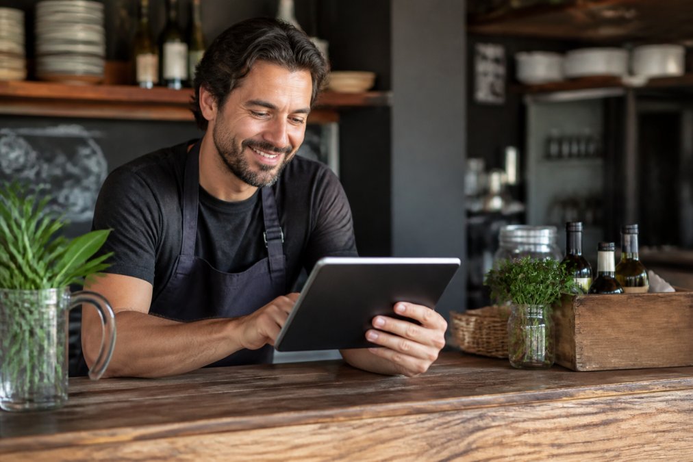 Dueño de restaurante pequeño usando una tablet en el mostrador, viendo una carta digital en la pantalla con código QR visible, sonriendo mientras trabaja