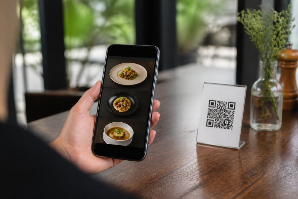 Mano de cliente sosteniendo un smartphone en una mesa de restaurante, escaneando código QR y viendo menú digital responsive en la pantalla