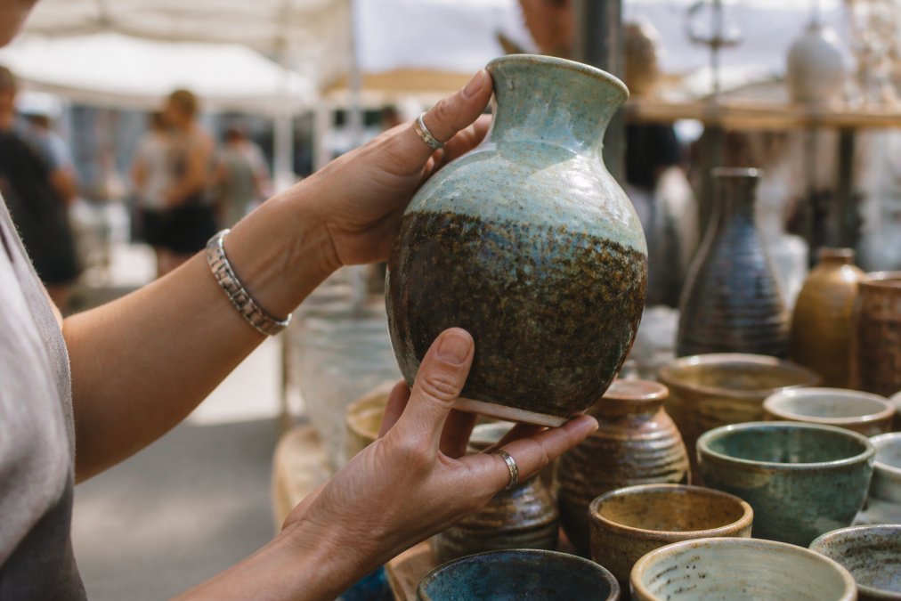 Close-up of hands examining a handmade ceramic vase at an artisan craft market, natural daylight illuminating the texture and glaze details