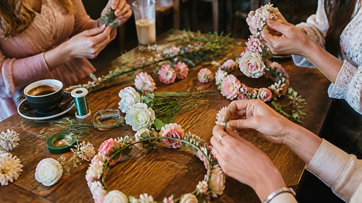 Close-up of guests' hands creating DIY flower crowns at a small café table with silk flowers, floral wire, and completed crowns visible in warm café lighting