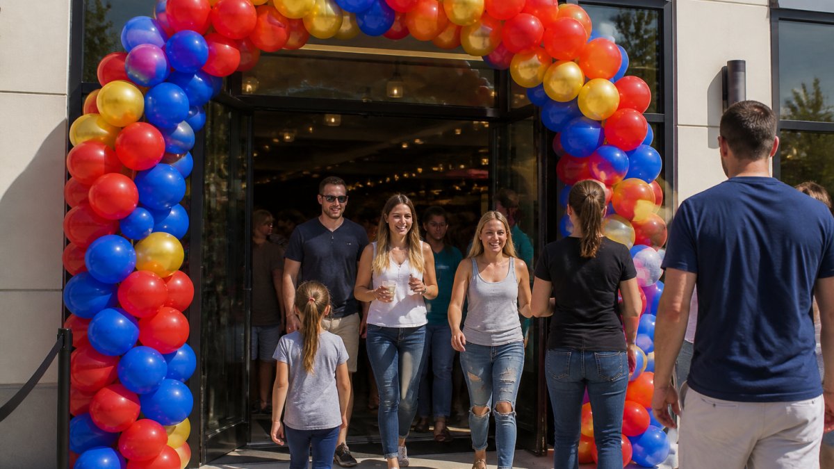 Colorful balloon arch framing a storefront entrance with red, blue, and gold balloons, customers smiling as they walk through the decorated doorway on a sunny day