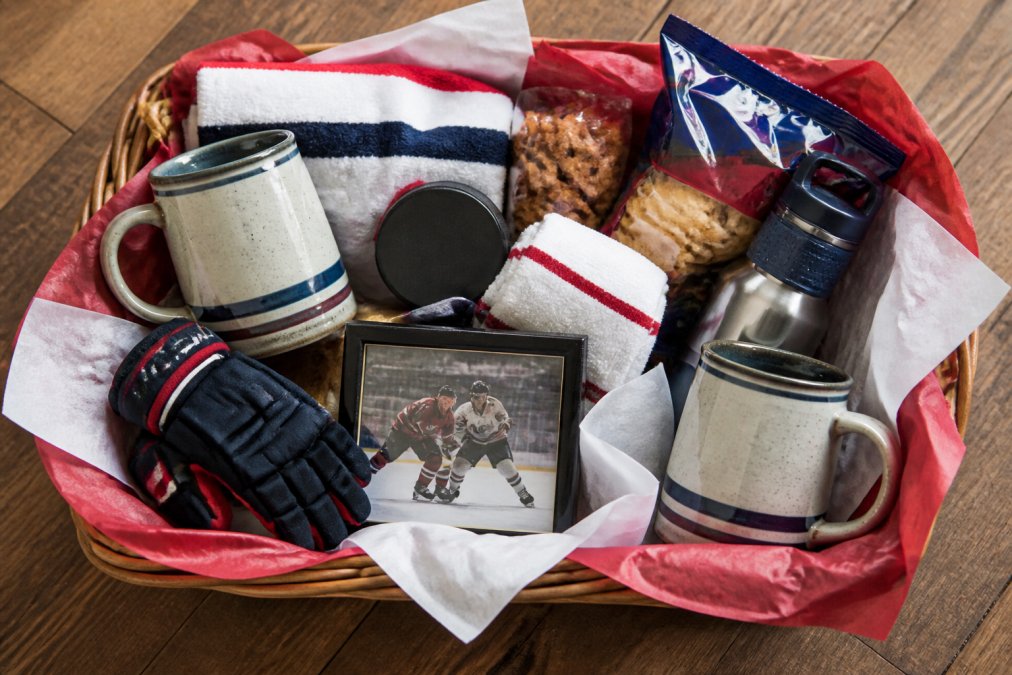 Overhead view of a beautifully arranged hockey gift basket with vintage ceramic mugs, microfiber towels, team-themed snacks, and memorabilia nestled in a woven basket lined with red and white tissue paper