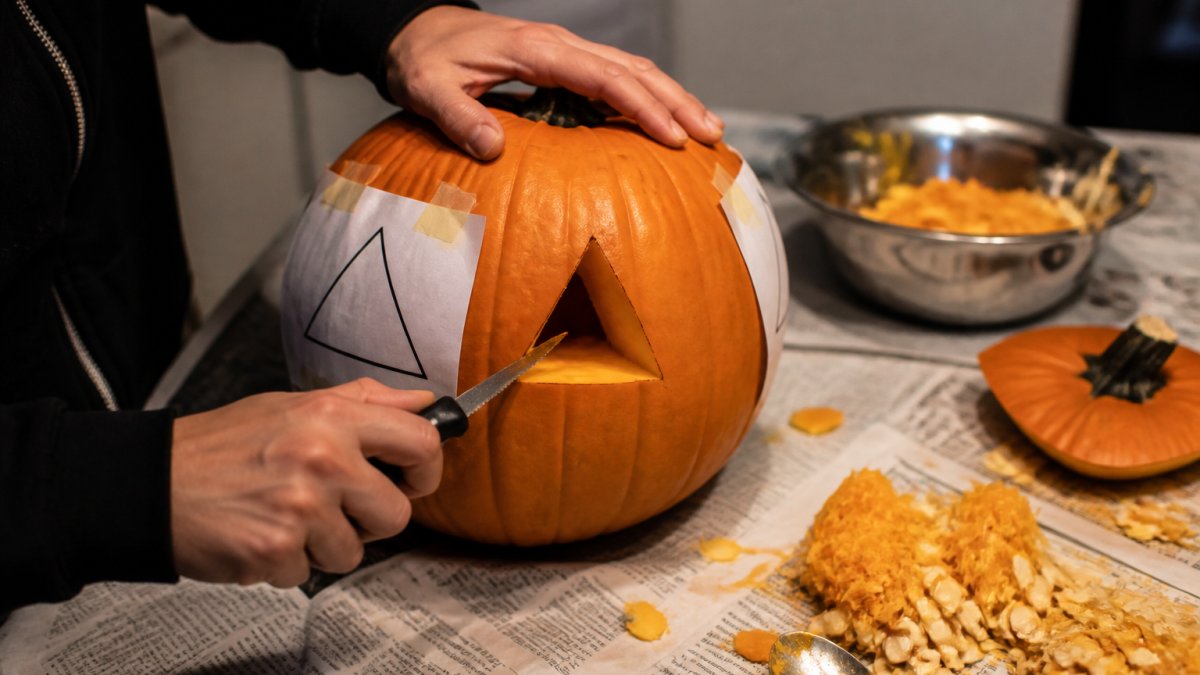 Close-up of hands using a small serrated knife to carve a simple triangle eye into a pumpkin on a newspaper-covered kitchen table, pumpkin pulp and seeds scattered nearby, a printed paper stencil taped to the gourd, warm overhead kitchen lighting