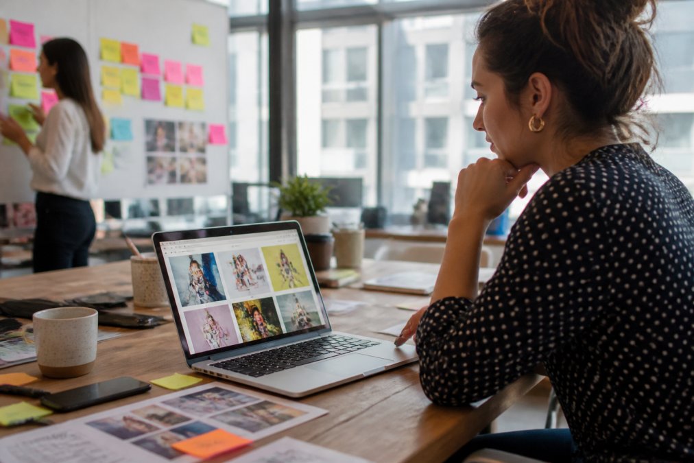 A marketing professional reviewing colorful social media ad creatives on a laptop screen in a bright modern office, with sticky notes and a coffee cup on the desk