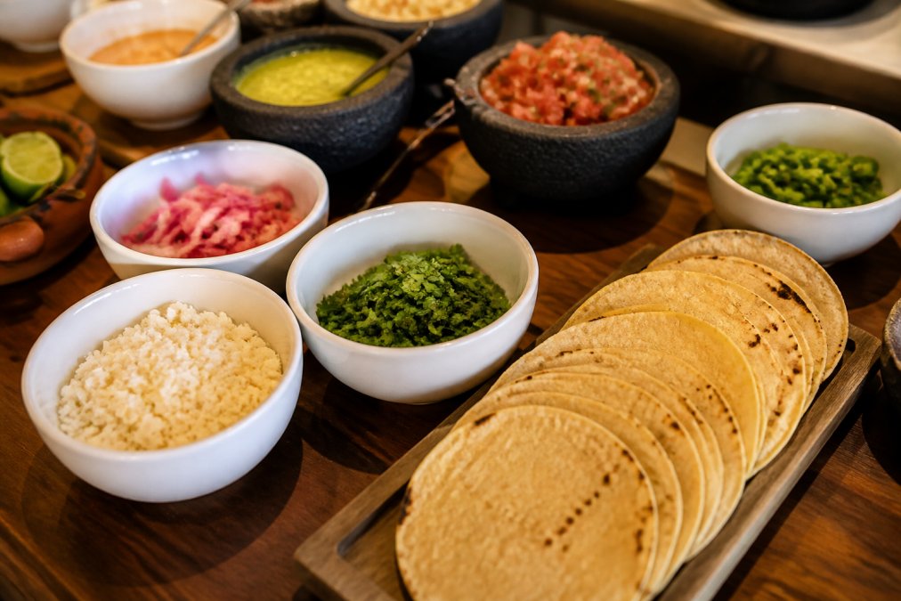 Elegantly arranged gourmet taco bar with small white bowls of cotija cheese, pickled red onions, and fresh cilantro, warm corn tortillas stacked on a wooden board, soft warm lighting on natural wood serving table