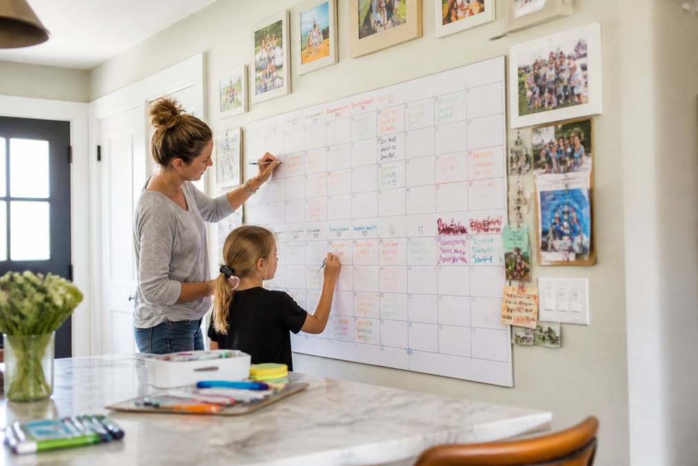 Bright kitchen wall displaying a large wall-sized calendar with colorful markers, family photos, and activity cards pinned nearby, with a parent and child adding notes together