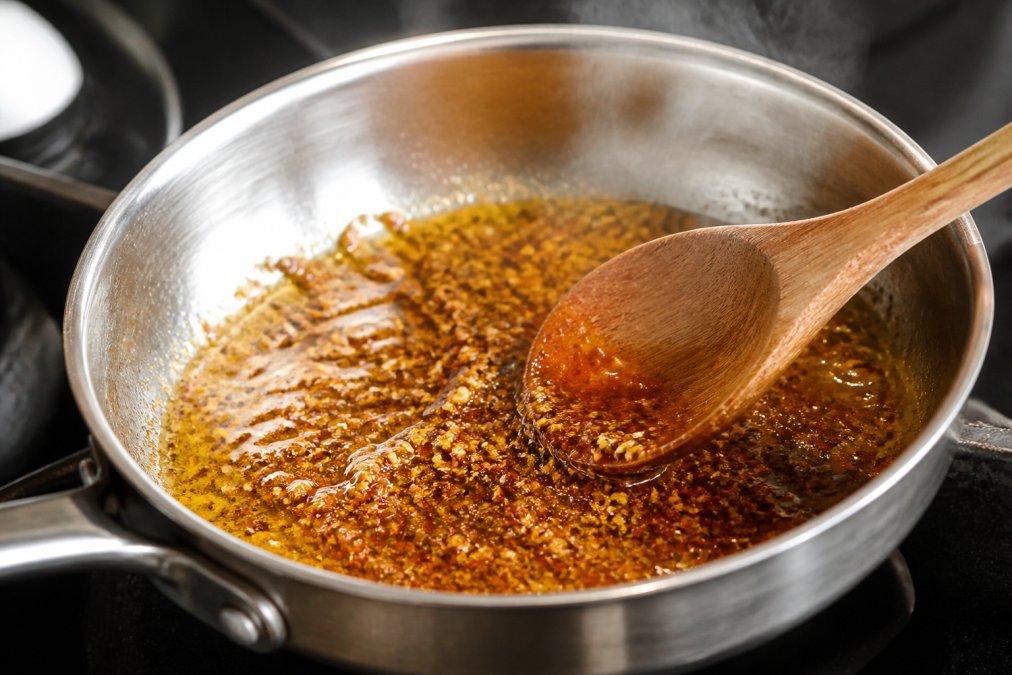 Close-up of vibrant spice blend being stirred in golden oil in a small stainless steel pan with steam rising, showing individual spice particles glistening in warm light