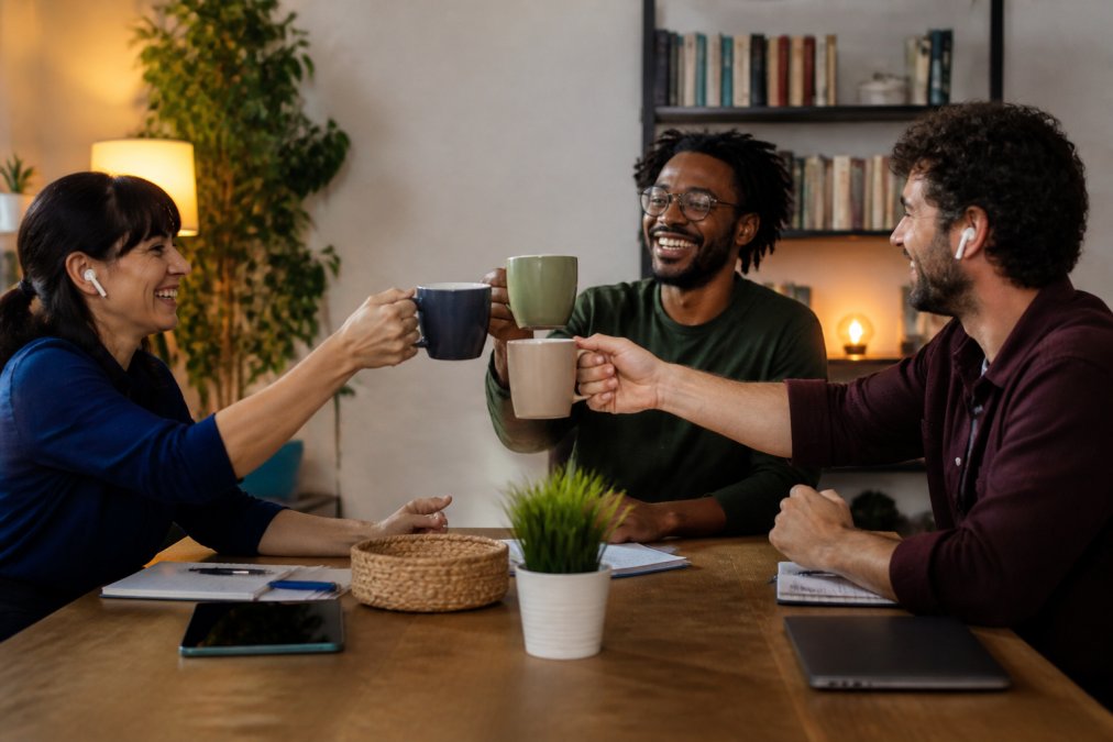 Remote employees on a video call smiling and raising coffee mugs in a toast to celebrate together from their home offices with warm lighting