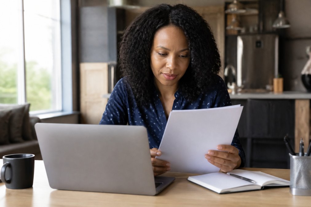 Professional woman reviewing her credit report on a laptop at home, sitting at a desk with a coffee cup and notebook beside her, natural window lighting