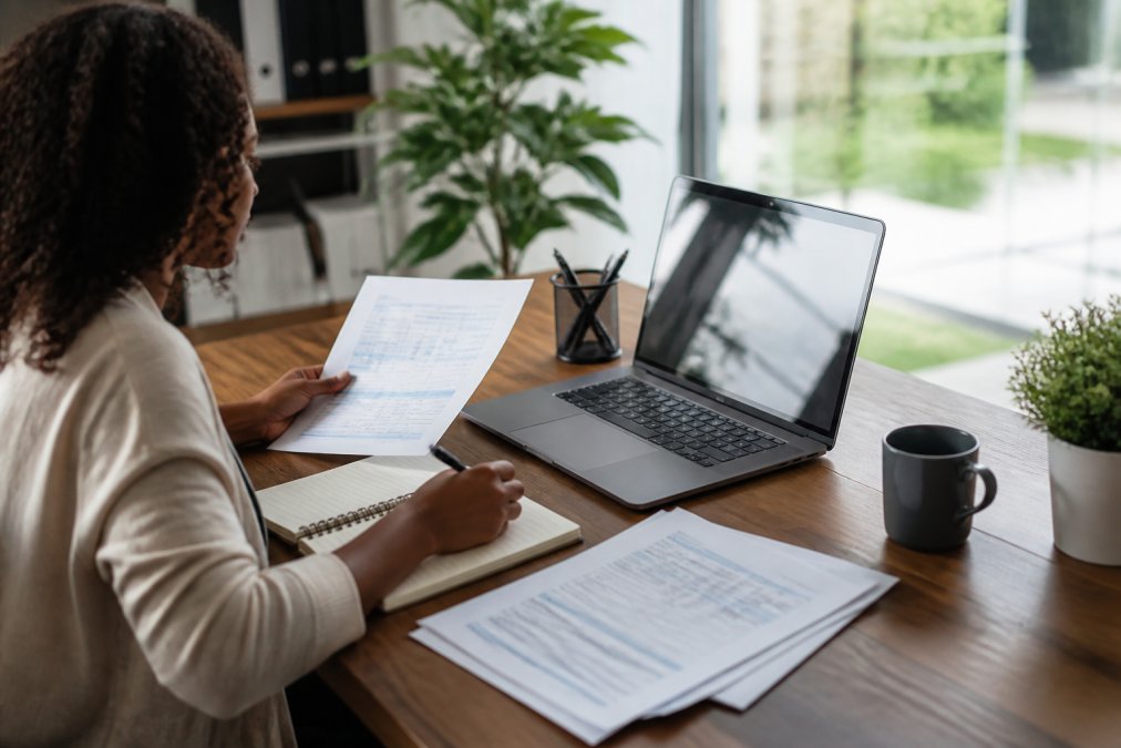 Person sitting at a desk reviewing printed credit report documents and taking notes while using a laptop displaying credit information, natural office lighting