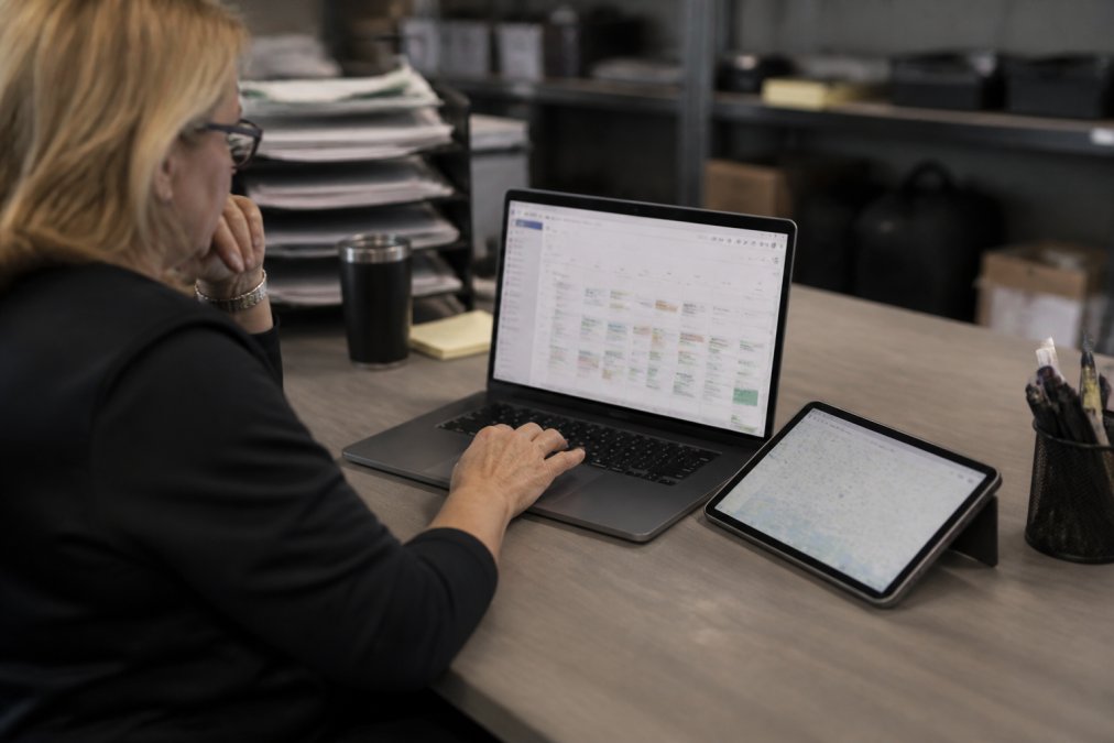 Office manager or dispatcher at a desk using a laptop to review crew schedules and work orders, with a tablet showing field team locations nearby